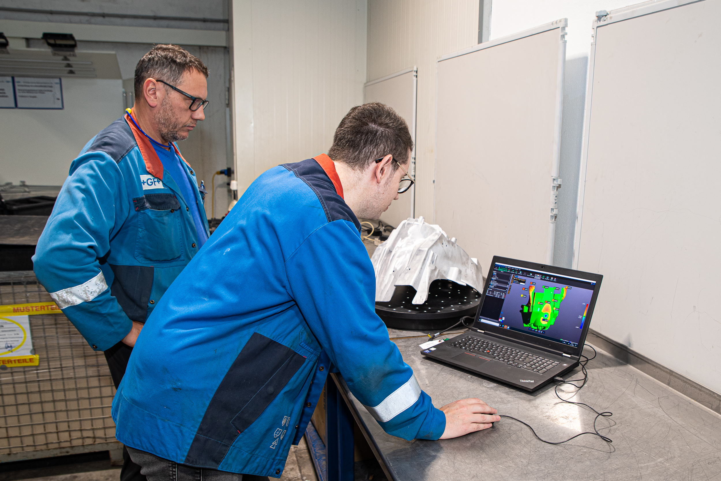 Two employees from GF Casting Solutions in blue GF Casting look at a scan of a part with colormap on a laptop. Next to it is a white component on a black turntable covered with dots