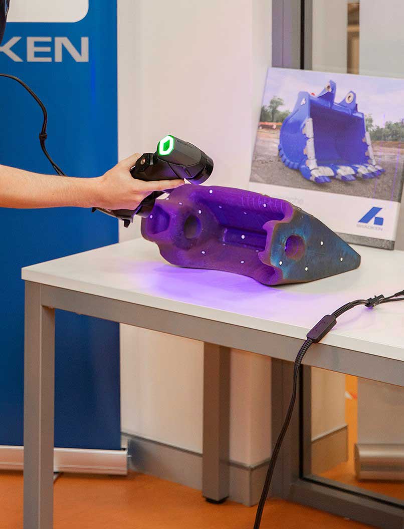 A worker at Bradken using the HandySCAN 3D scanner to scan an excavator bucket tooth on a table in front of a blue bucket picture.
