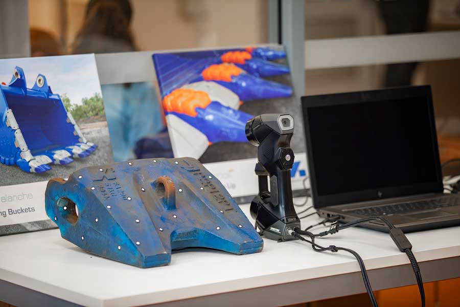 The HandySCAN 3D scanner is on a table between a blue escavator bucket tooth and a black laptop in front of pictures of the product.