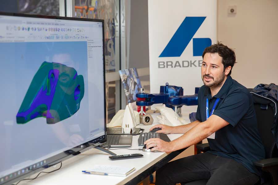 A Bradken employee sitting at a desk in front of a large screen displaying the model of an excavator bucket tooth in Scan-to-CAD Software. 