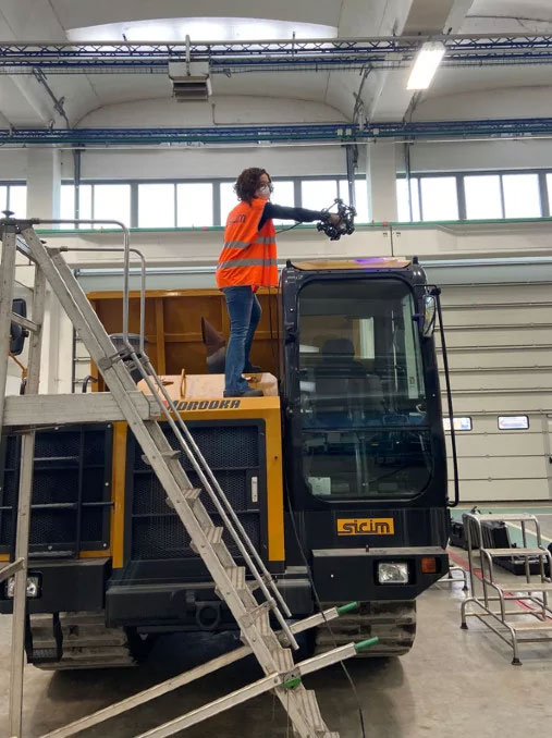 (Left) Woman standing on a yellow dump truck using a MetraSCAN 3D scanner to measure the driver cabin inside a warehouse. 