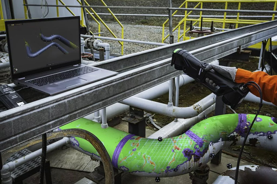 Man in a black polo shirt with yellow Zeppelin logo and black trousers scanning a yellow-colored engine block with the HandySCAN BLACK 3D scanner. On the right in the foreground is a laptop on a blue work trolley on which the scan can be seen
