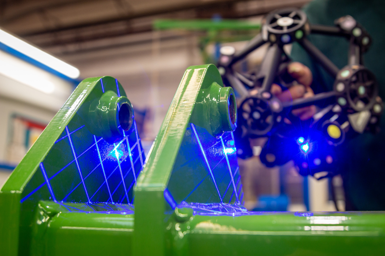 Un ingeniero sujeta un MetraSCAN 3D que emite líneas láser azules en una pieza grande con un acabado de superficie brillante.